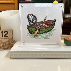 May include: A square acrylic frame containing a white fabric panel with an embroidered green rowboat, a brown wooden oar, a black fishing net, and a red tackle box. A small wooden sign reads 12 NORTH Embroidery, and a miniature green boat sits nearby.