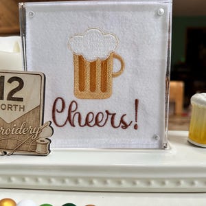 May include: A decorative display featuring a white felt square with an embroidered beer mug and the word "Cheers!". A small ceramic beer mug and a wooden sign with "12 North Embroidery" are also present.