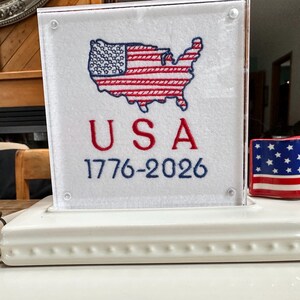 May include: A white ceramic display piece with a clear acrylic panel featuring an embroidered American flag map and the word "USA" with the years "1776-2026". A small ceramic flag is also present.