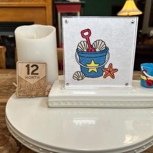 May include: A square white display piece featuring an embroidered blue bucket with a yellow star, a red shovel, and seashells. Beside it sits a white pillar candle, a small wooden sign reading 12 North Embroidery, and a miniature blue bucket.