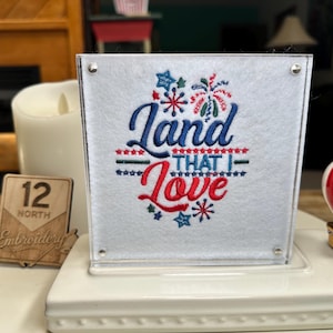 May include: A square white felt piece encased in a clear acrylic frame featuring embroidered text that reads Land That I Love in blue and red script. The design includes small red and blue star accents and firework motifs in red and blue.