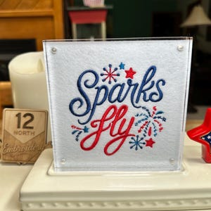 May include: A square white acrylic sign featuring the embroidered text Sparks fly in blue and red script. The design includes small red and blue firework motifs and stars. The sign is mounted on a white tiered base with a red star accent.