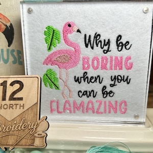 May include: A decorative display featuring a pink flamingo theme. It includes a framed embroidery with a flamingo and the text "Why be BORING when you can be FLAMAZING." Also, a wooden sign and a flamingo figurine.