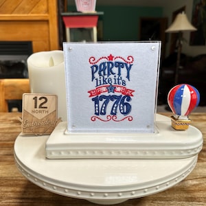 May include: A square white fabric panel featuring red and blue machine embroidery that reads Party like it's 1776. The panel is displayed in a clear acrylic frame on a white ceramic tiered stand, accompanied by a small hot air balloon figurine.