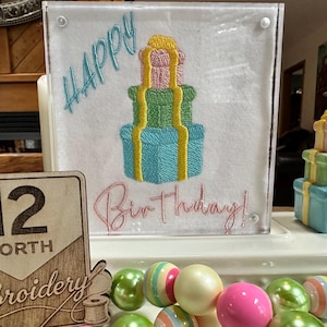 May include: A clear acrylic block with embroidered text reading "Happy Birthday!" and a stack of gifts. A wooden sign with the number 12 and the word "Embroidery" is in the foreground. Colourful beads are at the bottom.