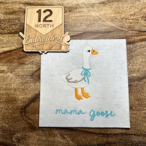 May include: A white felt square featuring an embroidered goose with an orange beak and feet, wearing a light blue ribbon around its neck. Below the goose, the text mama goose is embroidered in light blue. A wooden sign reads 12 NORTH Embroidery.