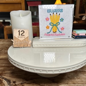 May include: A white ceramic pedestal stand holding a white LED candle, a wooden sign with the text 12 NORTH Embroidery, and a clear acrylic block featuring an embroidered design of yellow pencils, flowers, and the text ERASERS Made for Mistakes.