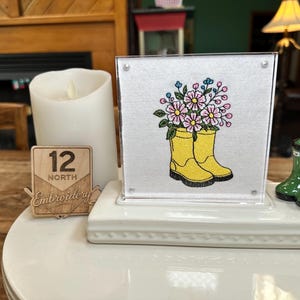 May include: A square white fabric panel featuring an embroidered design of yellow rain boots filled with pink and blue flowers, displayed in a clear acrylic frame on a white base, accompanied by a small wooden sign labeled 12 North Embroidery.