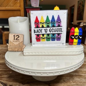 May include: A white ceramic tiered tray stand featuring a square sign with embroidered colorful crayons and the text BACK TO SCHOOL. Beside the sign are three small crayon-shaped figures in red, yellow, and blue, and a wooden 12 North Embroidery tag.
