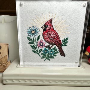 May include: A square acrylic frame containing white fabric with an embroidered red cardinal bird, blue and pink flowers, green leaves, and a yellow sunburst pattern. The frame sits on a white rectangular base with a beaded edge detail.