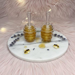 May include: Two clear glass bottles filled with shimmering gold liquid, resembling honey, sit on a marble tray. The bottles have a beehive design. The tray is decorated with lavender sprigs and small bee-shaped decorations.