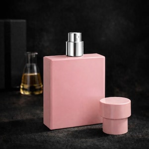 Puede incluir: Un frasco de perfume rectangular rosa con un pulverizador met&aacute;lico plateado y un tap&oacute;n cil&iacute;ndrico a juego. Un peque&ntilde;o frasco de vidrio con un l&iacute;quido amarillo est&aacute; en el fondo. Los objetos est&aacute;n sobre una superficie oscura.