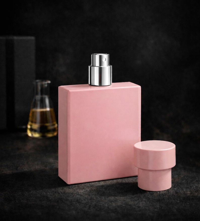Puede incluir: Un frasco de perfume rectangular rosa con un pulverizador met&aacute;lico plateado y un tap&oacute;n cil&iacute;ndrico a juego. Un peque&ntilde;o frasco de vidrio con un l&iacute;quido amarillo est&aacute; en el fondo. Los objetos est&aacute;n sobre una superficie oscura.