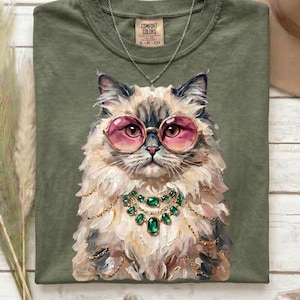May include: Olive green Comfort Colors t-shirt featuring a detailed illustration of a cat wearing pink sunglasses and a green gem necklace. The cat has fluffy fur and is centered on the shirt. A silver necklace is also visible.