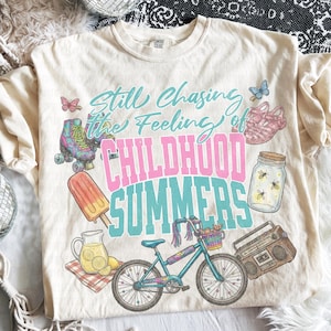 May include: Cream-colored t-shirt with a graphic design featuring the text "Still Chasing the Feeling of Childhood Summers." The design includes illustrations of a bicycle, roller skates, a popsicle, and a jar of fireflies.