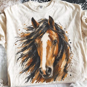 May include: A cream-colored t-shirt featuring a painterly graphic of a brown horse with a white blaze on its face and a flowing black mane. The artwork uses visible brushstrokes and includes scattered dark and tan paint splatters around the horse.