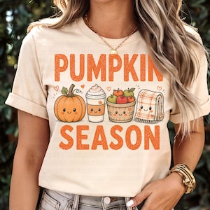 Pode incluir: Uma t-shirt cor de creme com o texto laranja PUMPKIN SEASON. O gráfico inclui uma abóbora sorridente, um copo de café com natas, um cesto de maçãs e uma manta aos quadrados dobrada, todos com caras amorosas e pequenos corações.