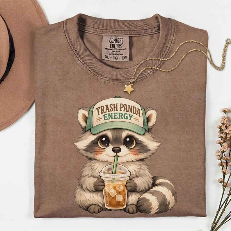 May include: A brown Comfort Colors t-shirt featuring a cartoon raccoon wearing a green hat with the text "Trash Panda Energy." The raccoon is holding a drink with a straw. A gold necklace with a star pendant is on the shirt.