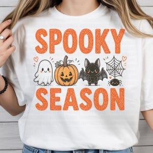 Könnte beinhalten: Ein weißes T-Shirt mit dem orangefarbenen Schriftzug SPOOKY SEASON und einer grafischen Illustration eines weißen Geistes, eines orangefarbenen Kürbisses, einer schwarzen Fledermaus und einer schwarzen Spinne an einem Netz, verziert mit kleinen orangefarbenen Herzen.