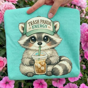 May include: A light teal t-shirt featuring a cartoon raccoon wearing a green hat with the text "Trash Panda Energy." The raccoon is drinking from a clear cup with ice cubes. The raccoon has big brown eyes and gray and white fur.