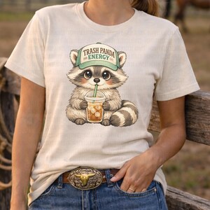 May include: Cream-colored t-shirt featuring a cartoon raccoon wearing a green hat with the text "Trash Panda Energy." The raccoon is holding a clear cup with iced coffee and a green straw. The shirt is paired with blue jeans and a brown belt.