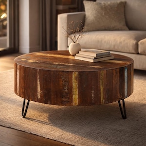 May include: A round coffee table with a rustic, reclaimed wood design. The tabletop features a mix of brown, yellow, and blue tones. The table is supported by black metal hairpin legs. Books and a small vase with dried flowers are on top.