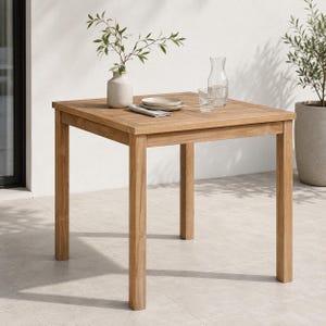 May include: A square wooden table with four straight legs and a slatted top surface. On the table sits a light-colored ceramic vase containing a green leafy branch, a stack of two white plates, a folded white napkin, and a clear glass carafe.