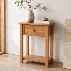 May include: A light brown wooden side table with a single drawer and a lower shelf. A vase with greenery, a small bowl, and books are on top. The table has a dark knob and is approximately 28 inches tall.