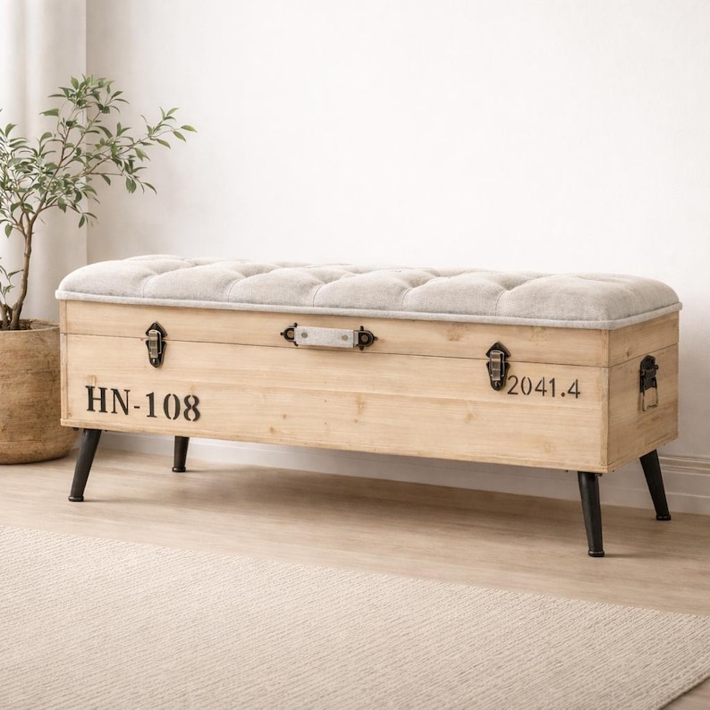 May include: A light wood storage bench with a tufted, light gray upholstered top. The bench has black metal legs and hardware. The front of the bench has the numbers "HN-108" and "2041.4" printed on it.