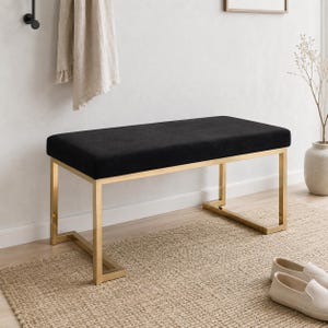 May include: A rectangular bench featuring a black upholstered seat cushion supported by a polished gold-tone metal frame with a geometric base design. The bench has a clean, structured silhouette with four vertical legs connected by horizontal bars.