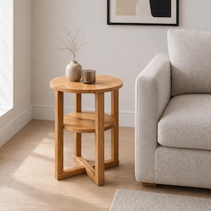 May include: A round, light-brown wooden side table with a lower shelf. A small vase with dried stems and a dark glass candle holder sit on top. The table is next to a light-colored sofa.