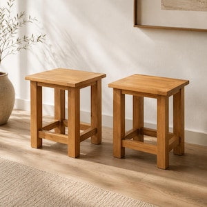 May include: Two square wooden stools with a natural finish. Each stool has a square seat and four sturdy legs connected by a lower horizontal support. The stools are made of light brown wood and are placed on a light-colored wooden floor.