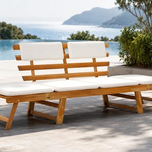 May include: A wooden outdoor bench with a light brown finish and white cushions. The bench has a slatted back and seat cushions. The bench is placed outdoors near a pool with a view of the ocean and mountains.
