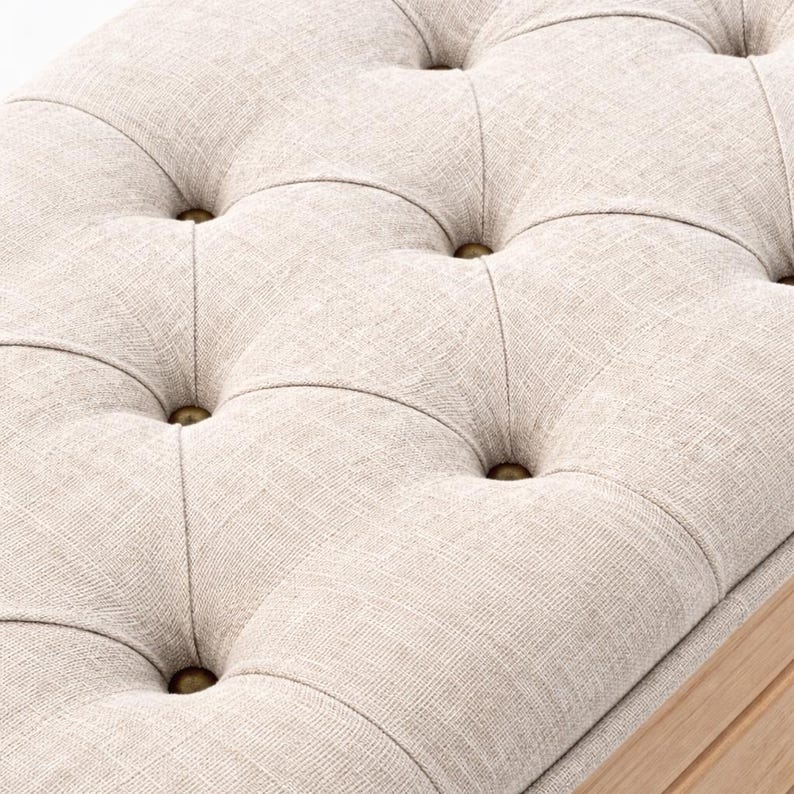 May include: Close-up of a beige upholstered bench with a tufted design. The fabric has a textured appearance, and the tufts are secured with brass-colored buttons. The bench has a light-colored wooden frame.