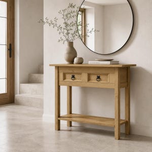May include: A light brown wooden console table featuring two drawers with black ring pulls and a lower shelf. The table is topped with a beige ceramic vase containing thin branches, a small round stone object, and a stack of books.