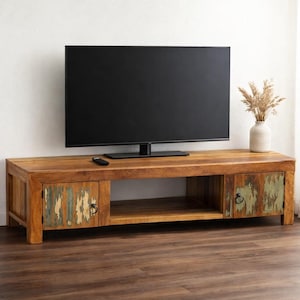 May include: A wooden TV stand with a natural finish, featuring two doors with distressed paint in shades of green and blue. The stand has an open shelf and a large TV on top. A vase with dried plants sits on the right.