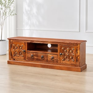 May include: A wooden TV stand with a warm brown finish. The stand features two doors with ornate carvings and brass accents, three drawers, and an open shelf. The doors have ring pulls, and the stand is on a wooden floor.