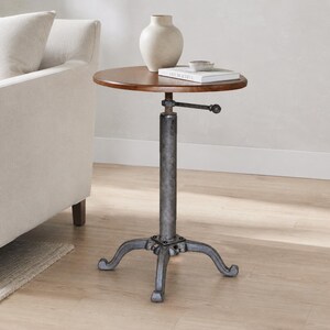 May include: A round wooden side table with a dark brown finish, featuring a central adjustable metal column with a hand crank mechanism and a three-legged cast iron base. A beige ceramic vase and a white book are placed on the tabletop.