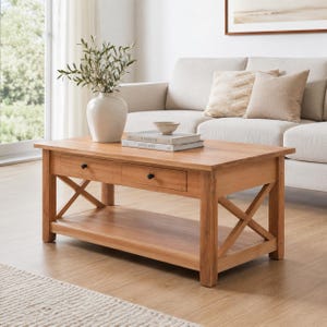 May include: A rectangular wooden coffee table with two drawers and an open shelf. The table has an X-shaped design on the sides and is made of light-colored wood. A white vase with greenery, books, and a bowl are on top.