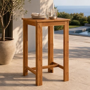 May include: A tall, square, light brown wooden bar table with four straight legs and a lower support crossbar. The tabletop features a stack of white plates, a folded white napkin, two clear drinking glasses, and a small white bowl.