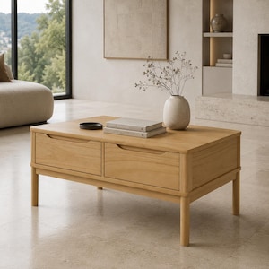 May include: A rectangular light wood coffee table featuring two front-facing drawers with integrated handle cutouts and four cylindrical legs. The tabletop holds a small dark circular dish, two stacked books, and a light-colored ceramic vase.
