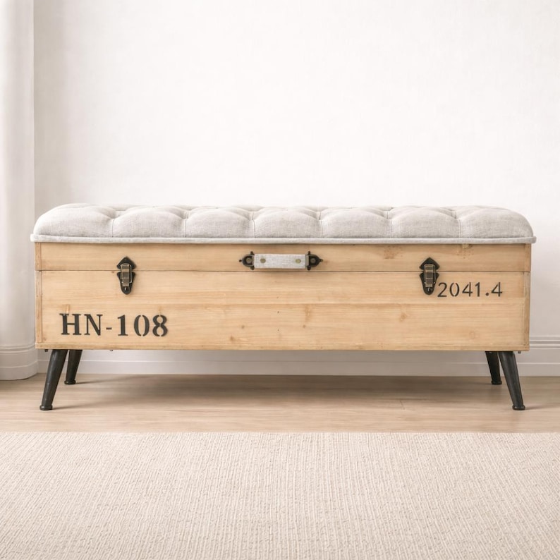 May include: A wooden storage bench with a tufted, light gray upholstered top. The bench has black metal legs and hardware. The front of the bench has the numbers "HN-108" and "2041.4" printed on it.