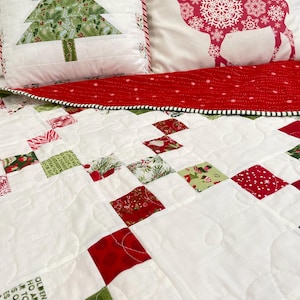 May include: A quilted bedding set featuring a white quilt with red and green patchwork squares, a white pillow with a green Christmas tree, and a white pillow with a red reindeer silhouette. The quilt has text printed on some fabric squares.