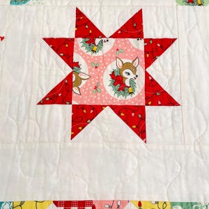 May include: A quilted fabric piece featuring a central eight-pointed star block with a pink background and a fawn illustration surrounded by a green wreath. The star is surrounded by white fabric with a wavy stitched pattern and holiday motifs.
