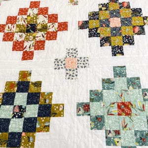 May include: A patchwork quilt featuring a white background with repeating cross-shaped blocks. The blocks are composed of smaller squares in various floral and botanical patterns, showcasing colours like rust orange, mustard yellow, navy blue, and sage green.
