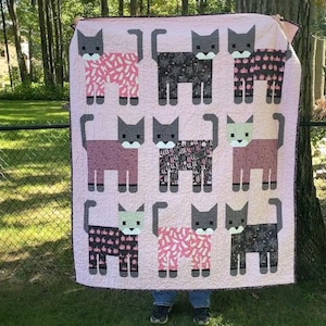 May include: A rectangular quilt featuring a light pink background with a grid of nine stylised cat figures. Each cat has a grey head and tail, with bodies patterned in various black, pink, and white designs including pumpkins, spiderwebs, and bones.