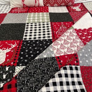 Peut inclure: Une courtepointe en patchwork composée de divers carrés de tissu sur le thème des fêtes en rouge, noir, blanc et gris. Les motifs incluent le tartan, des flocons, des silhouettes de rennes et le texte Have a Holly Jolly Christmas.