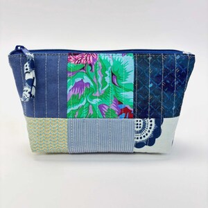 May include: A patchwork zippered pouch with a variety of fabrics in shades of blue, green, and floral patterns. The pouch has a blue zipper and a fabric pull. The base of the pouch is a light blue and yellow pattern.