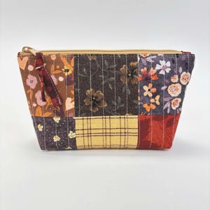 May include: A patchwork cosmetic bag with a gold zipper and a burgundy tassel. The bag is made of various fabric patterns, including floral, plaid, and solid colors in shades of brown, orange, and red. The bag is quilted.