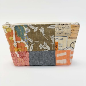 May include: A patchwork fabric pouch with a zipper closure. The pouch features a variety of patterned fabrics in shades of orange, blue, brown, and pink. The fabric includes floral and botanical prints, and text. A beaded pull is attached to the zipper.